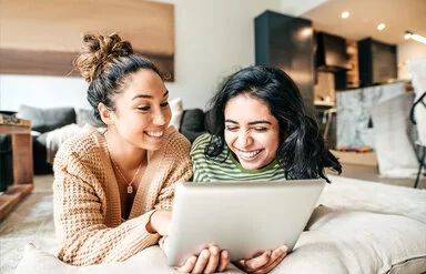 Zwei junge Frauen liegen gemütlich in einem Wohnzimmer auf dem Boden. Sie schauen sich etwas auf einem Tablet an, das sie amüsiert.