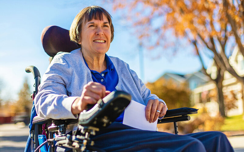 Eine Frau mit Multipler Sklerose fährt in einem Rollstuhl eine Straße entlang.
