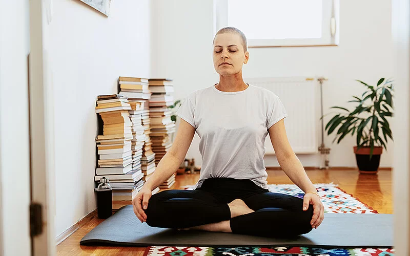 Eine weibliche Krebspatientin sitzt zuhause mit geschlossenen Augen im Schneidersitz auf einer Yogamatte.