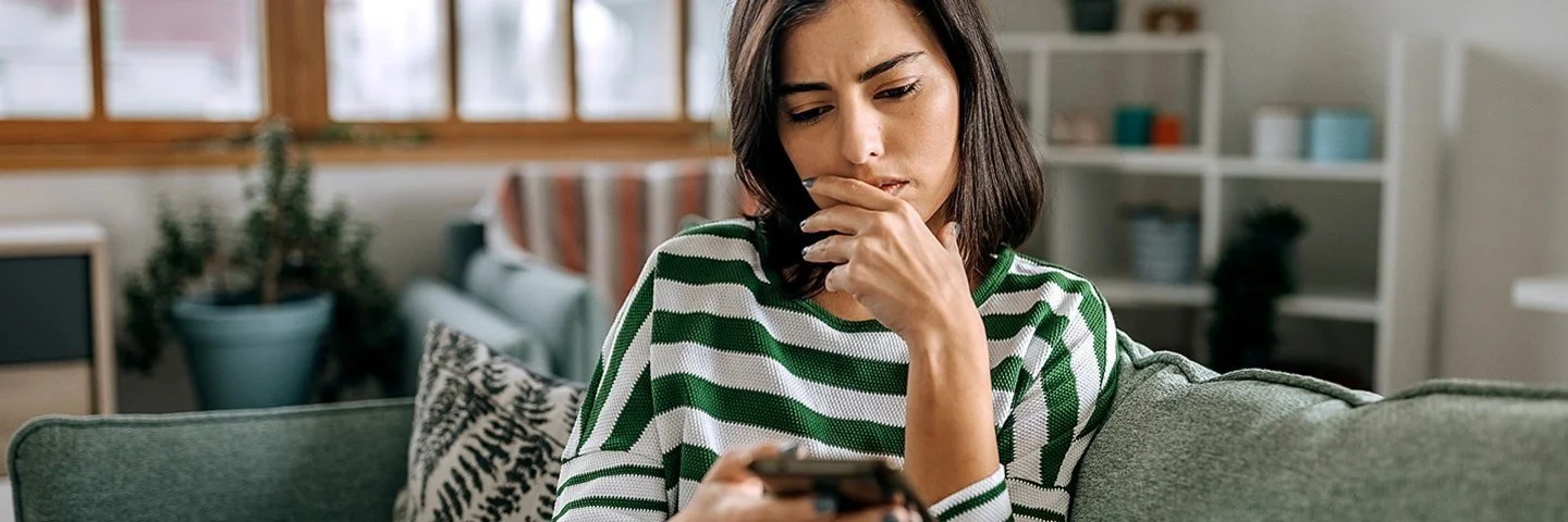Auf dem Sofa in einem Wohnzimmer sitzt eine junge Frau. Mit der rechten Hand hält sie ein Smartphone, mit der linken fasst sie sich ans Kinn. Sie blickt ernst auf das Display.