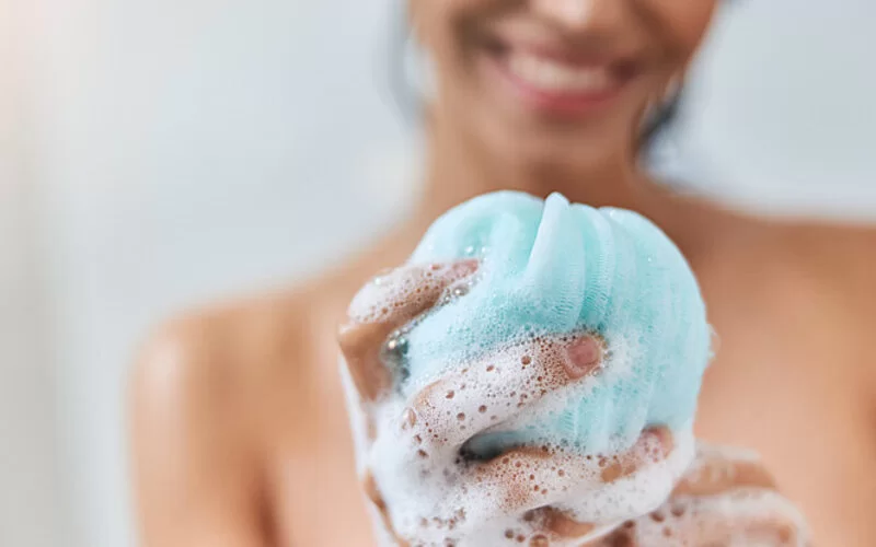 : Eine junge Frau hält bei ihrer täglichen Dusche einen schäumenden Schwamm in der Hand.