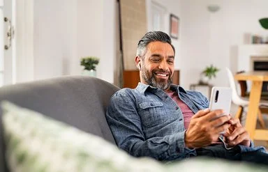 Ein Mann mittleren Alters sitzt entspannt auf dem Sofa und schaut zufrieden auf sein Smartphone.