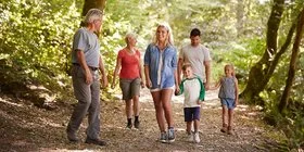 Eine Mehrgenerationenfamilie geht in einem Wald spazieren. Zu sehen sind zwei ältere Personen, ein mittelaltes Paar sowie zwei Kinder. Im Hintergrund sind Baumstämme und viele grüne Büsche zu erkennen.,