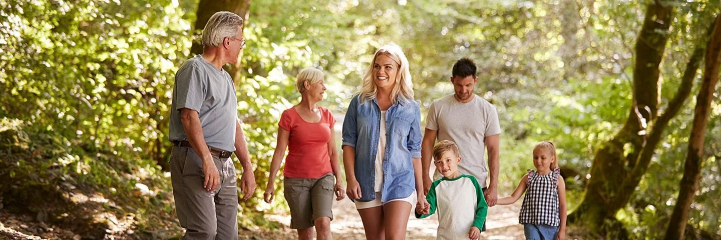 Eine Mehrgenerationenfamilie geht in einem Wald spazieren. Zu sehen sind zwei ältere Personen, ein mittelaltes Paar sowie zwei Kinder. Im Hintergrund sind Baumstämme und viele grüne Büsche zu erkennen.,