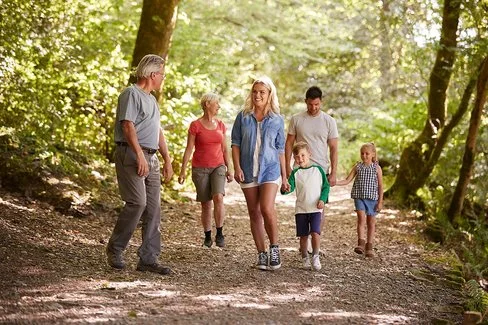 Eine Mehrgenerationenfamilie geht in einem Wald spazieren. Zu sehen sind zwei ältere Personen, ein mittelaltes Paar sowie zwei Kinder. Im Hintergrund sind Baumstämme und viele grüne Büsche zu erkennen., 