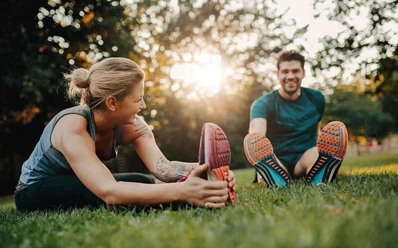 Ein Mann und eine Frau sitzen in Sportkleidung im Park auf Rasen. Sie stretchen sich und sind fröhlich.