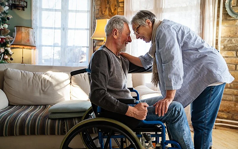 Ein älterer Herr sitzt im Rollstuhl in einem Wohnzimmer, eine Frau beugt sich über ihn. Die Beiden halten sich am Arm fest, schauen sich in die Augen und lächeln sich an.
