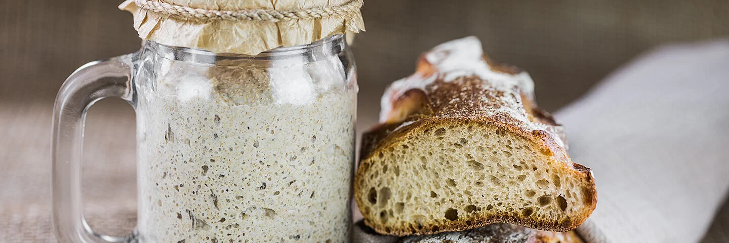 Links steht ein Glas gefüllt mit Sauerteig, rechts ein Sauerteigbrot.