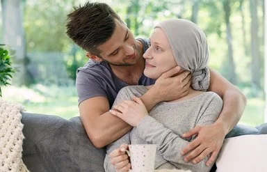 Ein Mann und eine Frau in einem Wohnzimmer in zärtlicher Umarmung. Die Frau trägt ein graues Kopftuch und hält eine Tasse in der Hand.