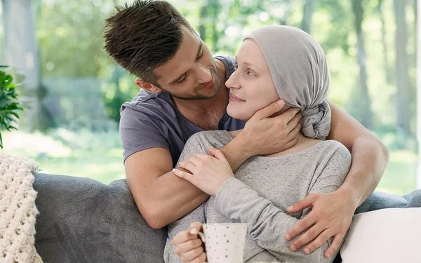 Ein Mann und eine Frau in einem Wohnzimmer in zärtlicher Umarmung. Die Frau trägt ein graues Kopftuch und hält eine Tasse in der Hand.