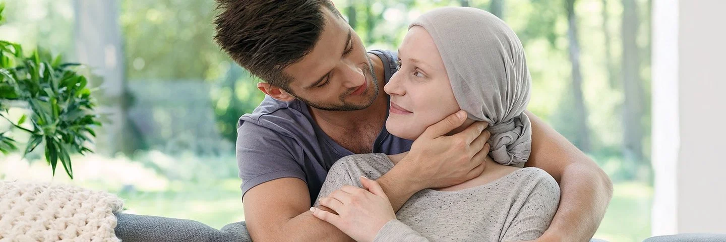 Ein Mann und eine Frau in einem Wohnzimmer in zärtlicher Umarmung. Die Frau trägt ein graues Kopftuch und hält eine Tasse in der Hand.