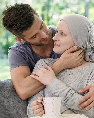 Ein Mann und eine Frau in einem Wohnzimmer in zärtlicher Umarmung. Die Frau trägt ein graues Kopftuch und hält eine Tasse in der Hand.