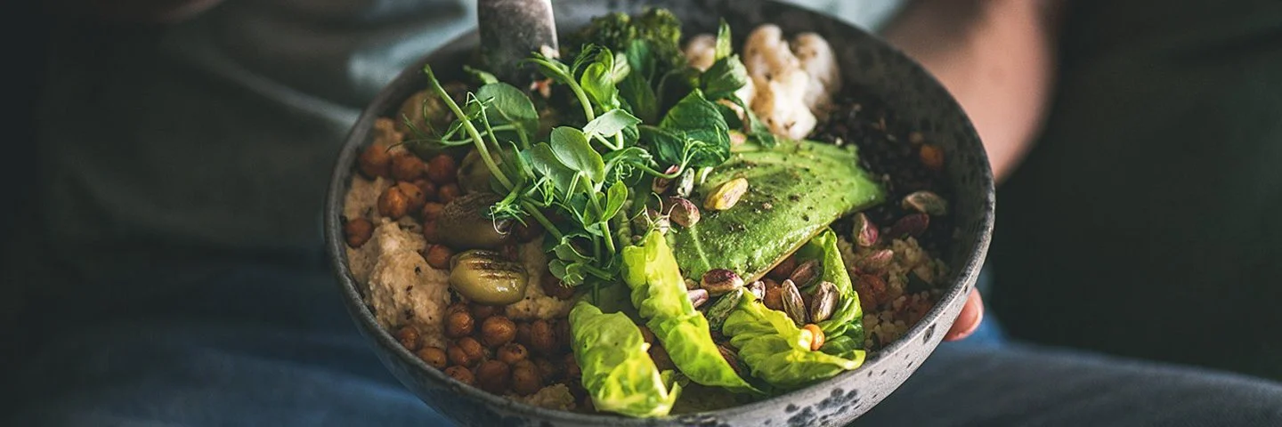 Eine nicht sichtbare Frau isst mit einer Gabel aus einer dunklen Keramikschüssel ein veganes Gericht aus Hummus, Salat, Bohnen, Blumenkohl und Avocado.