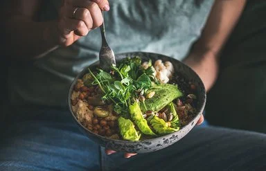 Eine nicht sichtbare Frau isst mit einer Gabel aus einer dunklen Keramikschüssel ein veganes Gericht aus Hummus, Salat, Bohnen, Blumenkohl und Avocado.