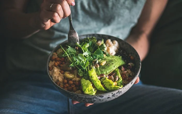 Eine nicht sichtbare Frau isst mit einer Gabel aus einer dunklen Keramikschüssel ein veganes Gericht aus Hummus, Salat, Bohnen, Blumenkohl und Avocado.