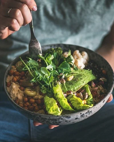 Eine nicht sichtbare Frau isst mit einer Gabel aus einer dunklen Keramikschüssel ein veganes Gericht aus Hummus, Salat, Bohnen, Blumenkohl und Avocado.