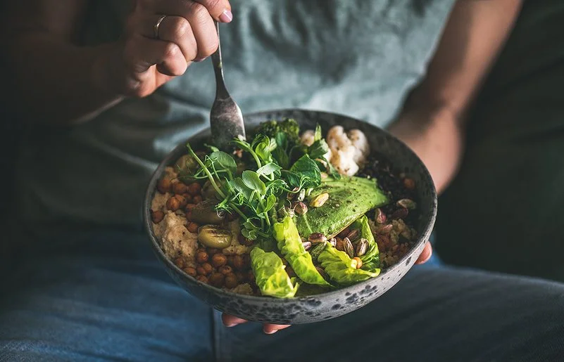 Eine nicht sichtbare Frau isst mit einer Gabel aus einer dunklen Keramikschüssel ein veganes Gericht aus Hummus, Salat, Bohnen, Blumenkohl und Avocado.