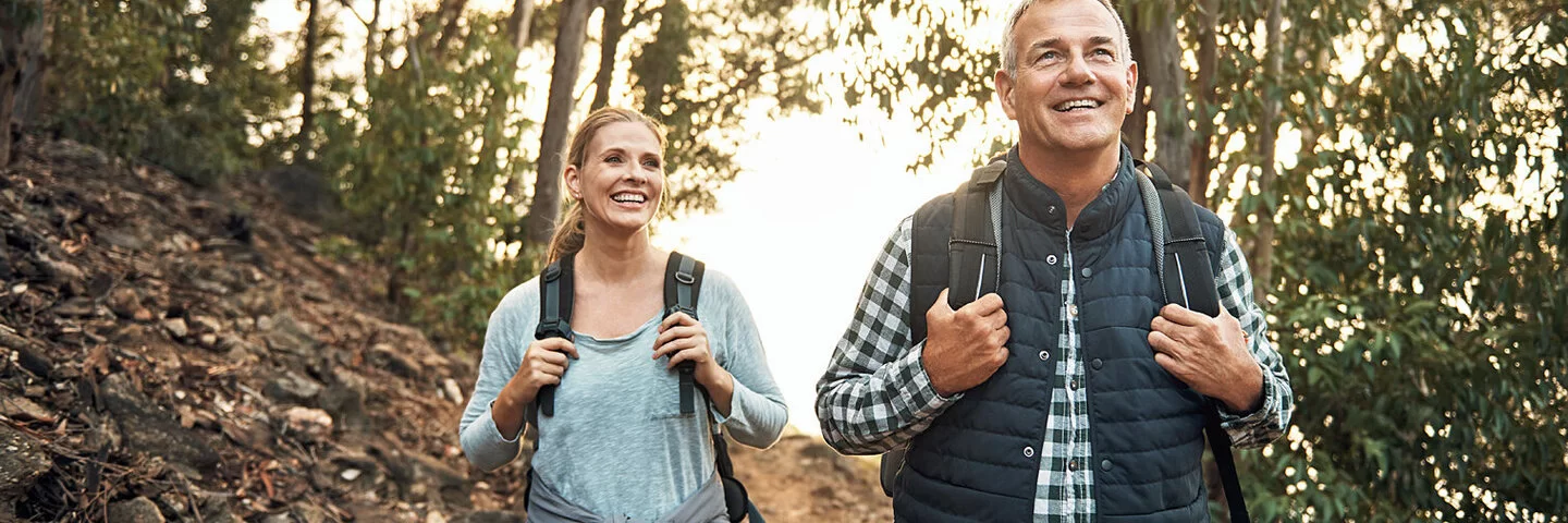 Ein Mann und eine Frau wandern mit Rucksäcken gut gelaunt an einem Waldhang.