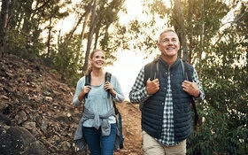 Ein Mann und eine Frau wandern mit Rucksäcken gut gelaunt an einem Waldhang.