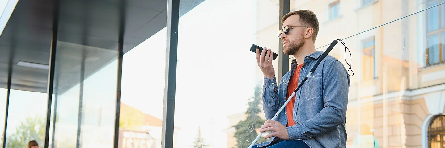 Ein junger Mann mit dunkler Sonnenbrille sitzt auf einer Bank. In der linken Hand hält er einen Blindenstock, in der rechten ein Smartphone.