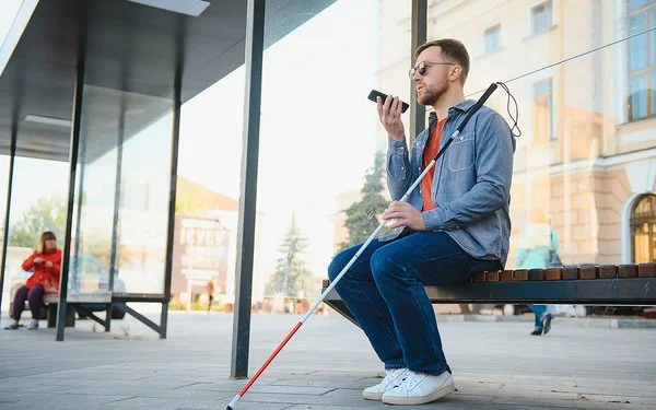Ein junger Mann mit dunkler Sonnenbrille sitzt auf einer Bank. In der linken Hand hält er einen Blindenstock, in der rechten ein Smartphone.