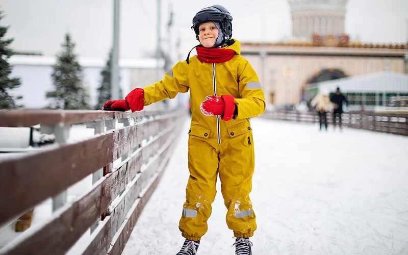 Junge mit gelbem Schneeanzug geht Schlittschuhlaufen.