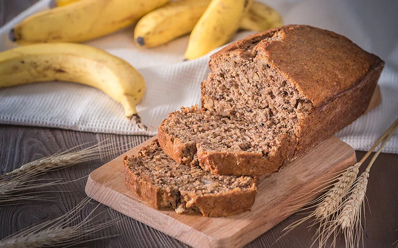 Ein frisch gebackenes Bananenbrot liegt auf einem Holzbrett. Daneben liegen reife Bananen und dekorative Weizenähren.