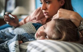 Ein Kind mit Lungenentzündung liegt auf der linken Seite im Bett, eine Frau berührt mit dem linken Arm seinen Kopf, während sie auf ein Fieberthermometer in der rechten Hand schaut.