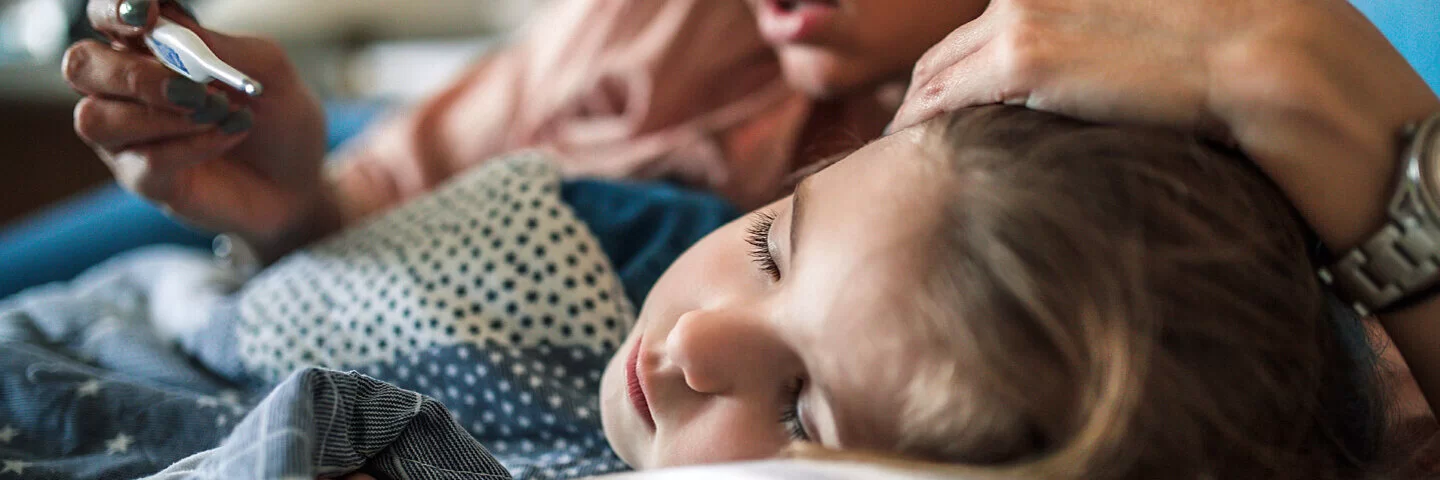 Ein Kind mit Lungenentzündung liegt auf der linken Seite im Bett, eine Frau berührt mit dem linken Arm seinen Kopf, während sie auf ein Fieberthermometer in der rechten Hand schaut.