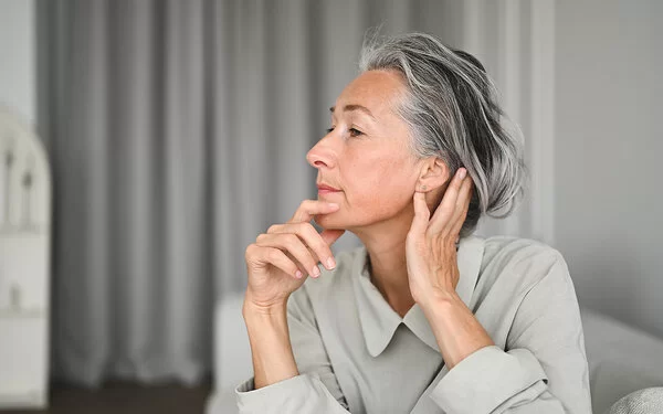 Eine Frau im seitlichen Portrait, die sich mit einer Hand an das Kinn und mit der anderen hinter das Ohr fasst.