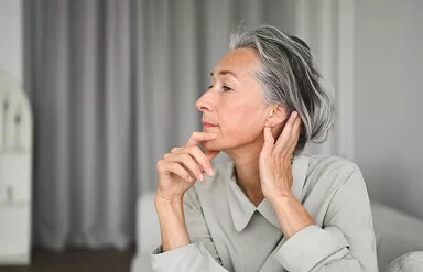 Eine Frau im seitlichen Portrait, die sich mit einer Hand an das Kinn und mit der anderen hinter das Ohr fasst.