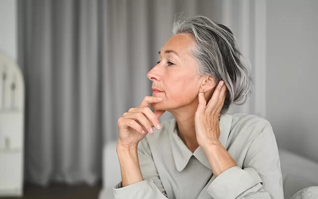 Eine Frau im seitlichen Portrait, die sich mit einer Hand an das Kinn und mit der anderen hinter das Ohr fasst.
