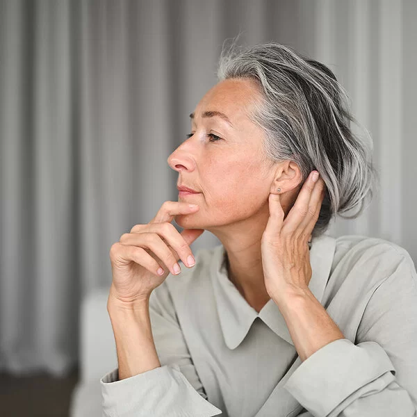 Eine Frau im seitlichen Portrait, die sich mit einer Hand an das Kinn und mit der anderen hinter das Ohr fasst.