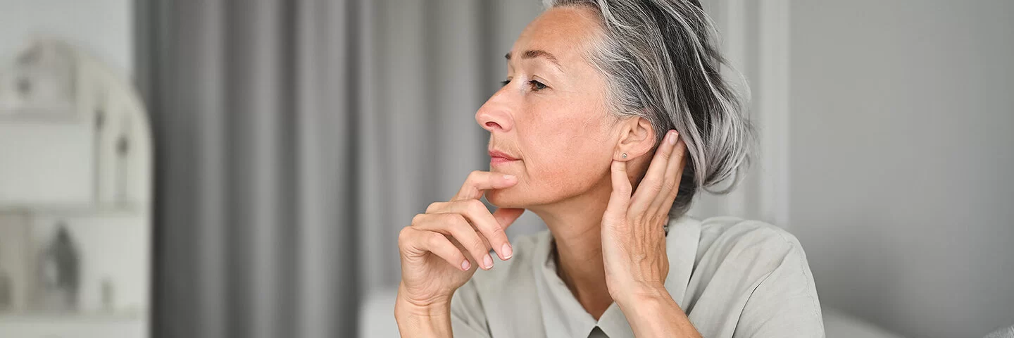 Eine Frau im seitlichen Portrait, die sich mit einer Hand an das Kinn und mit der anderen hinter das Ohr fasst.