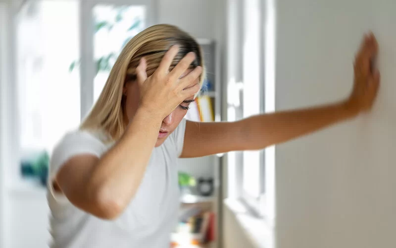 Eine junge Frau stützt sich während einer Schwindelattacke mit der rechten Hand an einer Zimmerwand ab, mit links greift sie sich an den Kopf.
