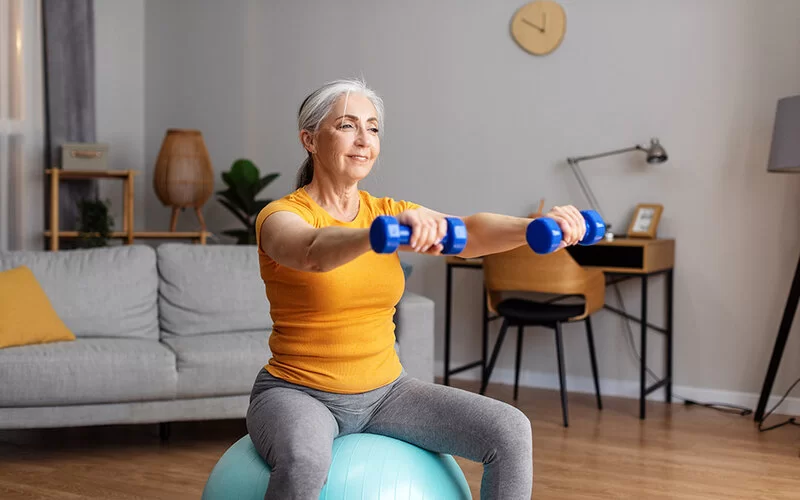Eina ältere Frau sitzt zu Hause auf einem Gymnastikball, die Arme nach vorne ausgestreckt, mit Kurzhanteln in den Händen, und lächelt dabei.