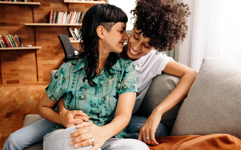 Ein queeres Paar sitzt eng umschlungen auf einem Sofa im Wohnzimmer.