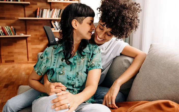 Ein queeres Paar sitzt eng umschlungen auf einem Sofa im Wohnzimmer.