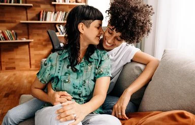 Ein queeres Paar sitzt eng umschlungen auf einem Sofa im Wohnzimmer.