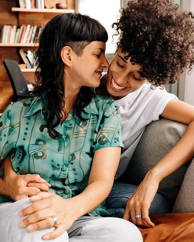 Ein queeres Paar sitzt eng umschlungen auf einem Sofa im Wohnzimmer.