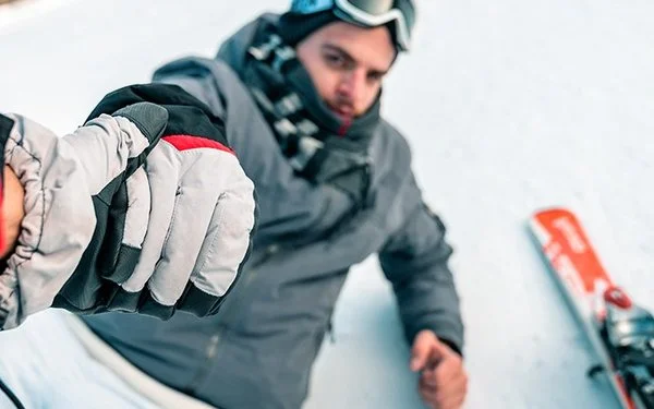 Eine Person mit Skibrille und Winterjacke sitzt auf dem Schnee und hält die Hand einer anderen Person, die einen rot-schwarzen Handschuh trägt. Neben der sitzenden Person liegt ein Ski. Die Szene spielt auf einer verschneiten Fläche.