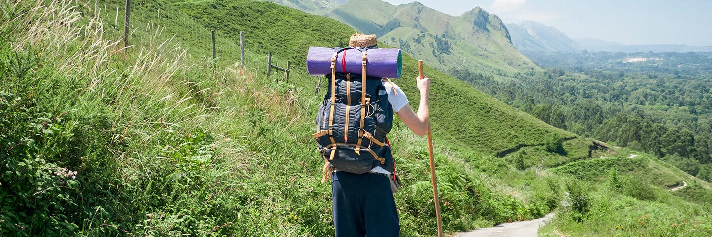 Ein junger Pilger mit Wanderstock läuft einen Pfad durch eine grüne Hügellandschaft entlang.