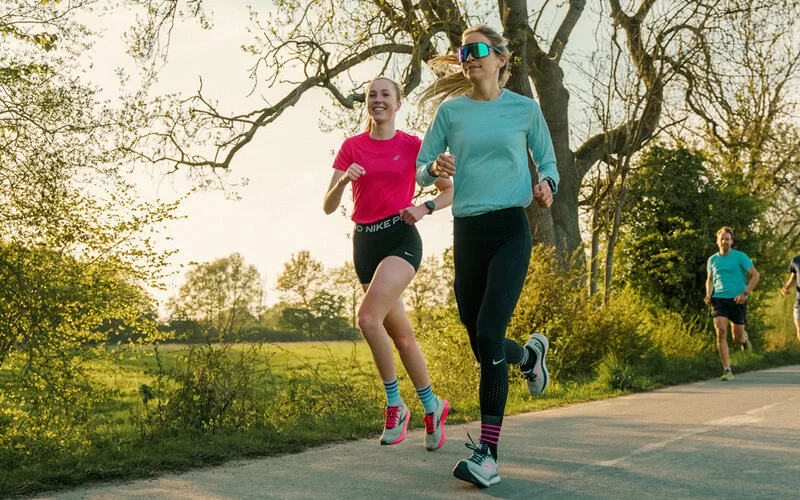 Zwei lächelnde Läuferinnen joggen einen Feldweg entlang während ein männlicher Läufer in der Distanz folgt.