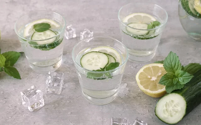 Drei Gläser mit Wasser, Zitronenscheiben, Gurkenscheiben und Minze auf einem grauen Untergrund. Links daneben liegen Eiswürfel, Minze und Zitrone, rechts zudem eine Salatgurke.