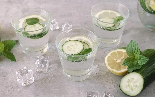 Drei Gläser mit Wasser, Zitronenscheiben, Gurkenscheiben und Minze auf einem grauen Untergrund. Links daneben liegen Eiswürfel, Minze und Zitrone, rechts zudem eine Salatgurke.