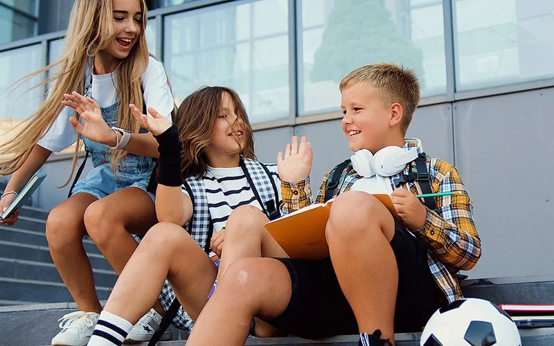 Drei Kinder sitzen und stehen auf einer Treppe vor einem Schulgebäude, zwei geben sich freudig Highfive, eines hält ein Buch und hat Kopfhörer um den Hals.