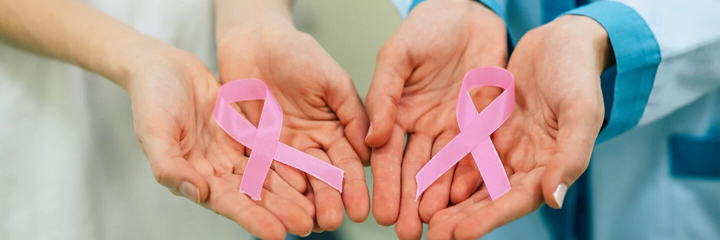 Zwei Frauen halten eine rosa Schleife in der Hand. Das Symbol steht für die Solidarität mit Frauen, die an Brustkrebs erkrankt sind oder ein höheres Brustkrebsrisiko haben.