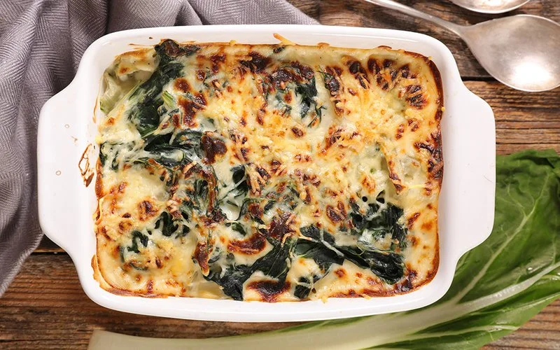 Mangold zubereitet: Die Spinatalternative als Auflauf mit Käse gratiniert. Daneben eine Gabel und ein Stiel des Gemüses.