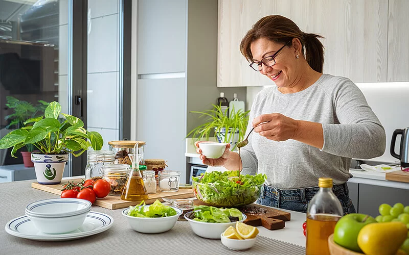 Eine Frau mittleren Alters bereitet in der Küche lächelnd einen Salat zu. Vor ihr stehen Schüsseln mit frischen Zutaten.