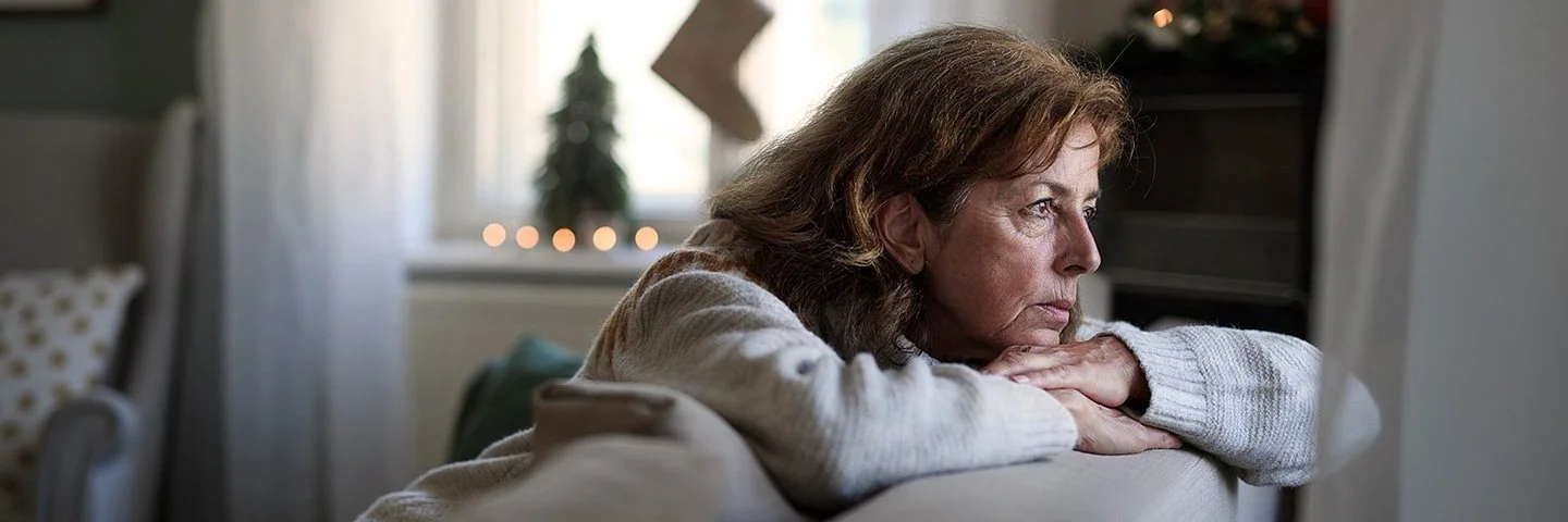 Eine ältere Frau sitzt auf dem Sofa und schaut nachdenklich aus dem Fenster. Im Hintergrund ist Weihnachtsdekoration zu sehen.
